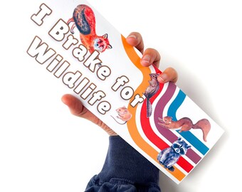 May include: A white sign with the text "I Brake for Wildlife" in brown lettering. The sign has a rainbow stripe design and features illustrations of a fox, squirrel, raccoon, chipmunk, and mouse.