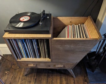May include: A wooden record player stand with a turntable on top and a drawer below. The stand has a built-in storage compartment for records. The stand is made of light brown wood and has a modern design.