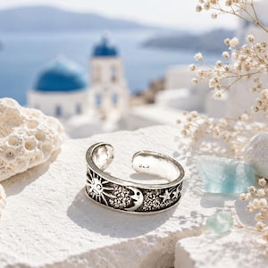 Könnte beinhalten: Ein silberfarbener, offener Ring mit einem geprägten Design aus Sonne, Mondsichel und Sternen. Auf der Innenseite des Rings ist deutlich der Text 925 eingeprägt, was auf die Metallzusammensetzung hinweist. Der Ring hat eine polierte Oberfläche.