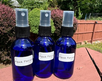 Pode incluir: Três frascos de spray azul cobalto com bicos pretos e tampas transparentes. Cada frasco tem um rótulo branco com as palavras "Agua Florida" em escrita preta. Os frascos são preenchidos com um líquido azul.