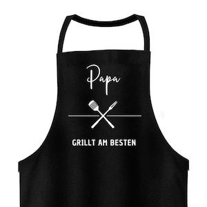 Könnte beinhalten: Schwarze Schürze mit dem Wort "Papa" in Schreibschrift über einer gekreuzten Gabel und einem Spatel. Darunter steht in Weiß der Text "Grillt am Besten".