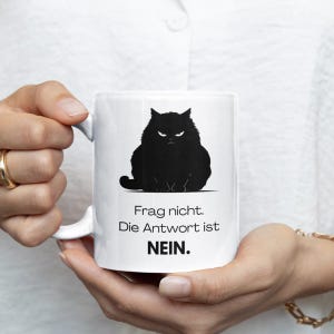 Könnte beinhalten: Weißer Keramikbecher mit einer schwarzen Silhouette einer grimmigen Katze und dem Text "Frag nicht. Die Antwort ist NEIN." Der Becher hat einen gebogenen Henkel und wird von einer Person gehalten.