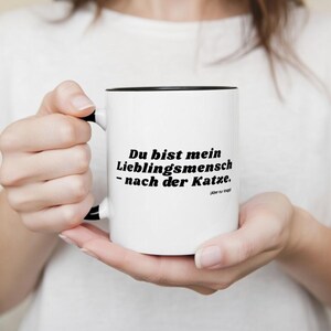 Könnte beinhalten: Weißer Keramikbecher mit schwarzem Innenraum und Henkel, gehalten von einer Person. Der Becher trägt den schwarzen Text "Du bist mein Lieblingsmensch - nach der Katze." Eine humorvolle Geschenkidee.