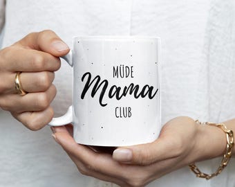 May include: White ceramic mug with the words "MÜDE Mama CLUB" in black script and sans-serif font. The mug has a white handle and is held by a person wearing a gold ring and bracelet.