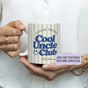 Könnte beinhalten: Weißer Keramikbecher mit vertikalen beigefarbenen Streifen. Der Becher zeigt den Text "OFFICIAL MEMBER Cool Uncle Club" in Blau, mit einem lächelnden Gesicht und dem Jahr 2026. Der Becher wird von einer Person gehalten.