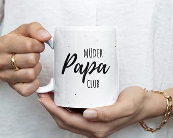 May include: White ceramic mug with the words "MÜDER Papa CLUB" in black script. The mug has a handle and is decorated with small black dots. The mug is held by a person wearing a gold ring and bracelet.