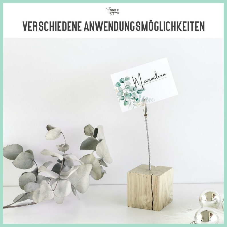 May include: A wooden base with a metal wire and clip holds a white card with green foliage and the name "Maximilian" written in black. The card is used as a place card or table number holder.