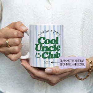 Könnte beinhalten: Eine weiße Keramiktasse mit blauen vertikalen Streifen. Die Tasse trägt den grünen Text "OFFICIAL MEMBER Cool Uncle Club" mit einem Smiley und dem Jahr 2026. Die Tasse wird von einer Person gehalten.