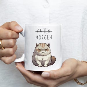Könnte beinhalten: Weiße Keramik-Tasse mit einem grimmigen Cartoon-Kater und dem Text "Guten Morgen". Die Katze hat einen strengen Gesichtsausdruck und braunes und graues Fell. Die Tasse hat einen Henkel.