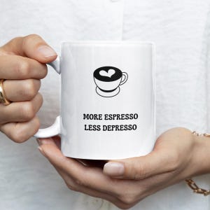 Könnte beinhalten: Weiße Keramik-Kaffeetasse mit einem schwarzen Kaffeetassen-Design und dem Text "MORE ESPRESSO LESS DEPRESSO". Die Tasse hat einen Henkel und wird von einer Person gehalten.
