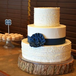 May include: A three-tiered wedding cake with white frosting and ruffled edges. The bottom tier is decorated with a navy blue ribbon and a large blue flower. The cake is sitting on a rustic wooden cake stand.