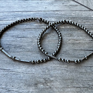 May include: Two black beaded bracelets with silver metal accents. The bracelets are made of small round beads and are lying on a wooden surface.