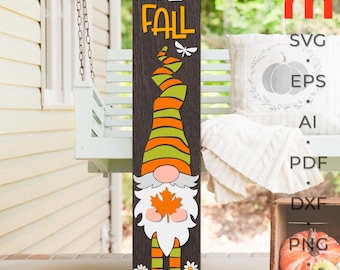 May include: A wooden sign with the words "Hello Fall" in orange and white letters. A gnome with an orange and green hat and a white beard is holding a leaf. The sign is on a porch with a swing.