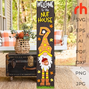 Peut inclure: Panneau en bois avec le texte "WELCOME to the NUT HOUSE" et une illustration de gnome. Le gnome porte un chapeau orange à pois jaunes et tient un gland. Le panneau est décoré de feuilles d'automne et est placé sur un porche.