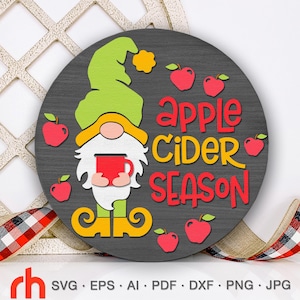 May include: A round, gray wooden sign with a gnome holding a red mug, surrounded by red apples. The words "Apple Cider Season" are in red. The gnome has a green hat and yellow shoes. A red and black plaid ribbon is in the background.