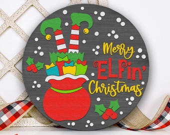 May include: A round, wooden Christmas sign with a gray background. It features a red sack filled with gifts, elf legs, and the text "Merry Elfin' Christmas" in yellow and red. Holly and white dots add festive detail.