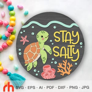 Op de afbeelding: Een ronde, donkergrijze houten plank met een cartoon schildpad, koraal en de woorden "STAY SALTY" in het geel. De schildpad is groen en oranje, en het koraal is roze en oranje. Bubbels en een zeester zijn ook aanwezig.