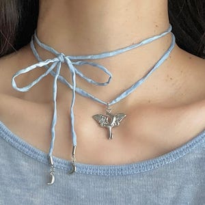 May include: A light blue choker necklace with a silver pendant. The necklace features a double-strand design tied in a bow at the front. A silver charm with a crescent moon and a silver pendant with an insect design are attached.