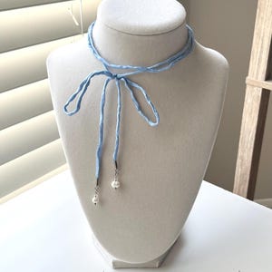 May include: A light blue ribbon choker necklace featuring a delicate tie-front design. The two ends of the ribbon are finished with silver-toned metal caps, each holding a small, round, white pearl bead that hangs down from the center knot.