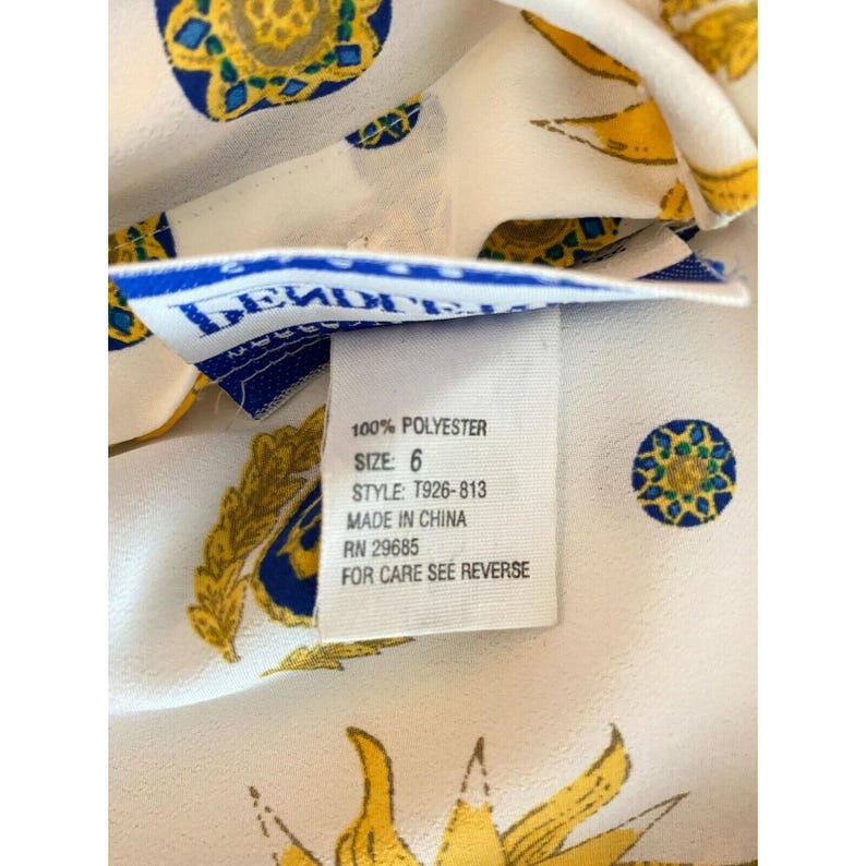 Puede incluir: Primer plano de una tela blanca con un patr&oacute;n repetido de dise&ntilde;os solares amarillos y azules. Una etiqueta con texto que incluye "100% POLYESTER" y "SIZE: 6" es visible. La etiqueta tambi&eacute;n incluye "MADE IN CHINA".