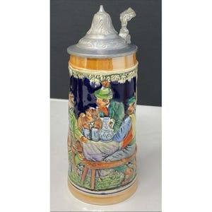 May include: A ceramic beer stein with a detailed relief scene of people at a table, featuring a pewter lid and handle. The stein has a cylindrical shape with a base and a decorative band around the top.