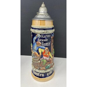 May include: A ceramic beer stein with a detailed relief scene of figures in traditional clothing. The stein has a blue background with the words "Kartenspielende Bauern!" and "Hopfen gibt". It features a metal lid and handle.