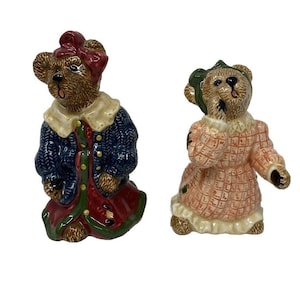 May include: Two ceramic bear figurines dressed in clothing. The bear on the left is wearing a blue sweater and a red and green dress. The bear on the right is wearing a pink and white dress.