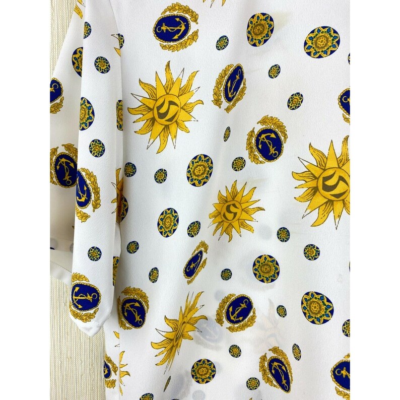Puede incluir: Tela blanca con un patr&oacute;n repetido de soles amarillos, anclas azules y dise&ntilde;os circulares dorados. La tela tiene un ligero brillo y parece ser un material ligero, posiblemente para una blusa o un vestido. El dise&ntilde;o es una mezcla de elementos n&aacute;uticos y celestiales.