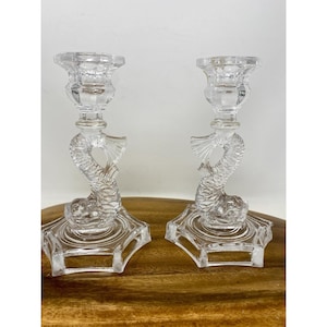May include: Two clear glass candle holders. Each holder features a dolphin design as the stem, with a hexagonal base and a cup for the candle. The candle holders are displayed on a wooden surface.