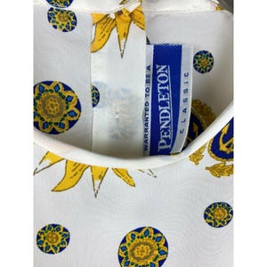 Puede incluir: Tela blanca con un estampado repetido de girasoles amarillos y dise&ntilde;os circulares azules y amarillos. Se ve una etiqueta Pendleton Classic azul y blanca. La tela parece ser parte de una camisa o blusa.