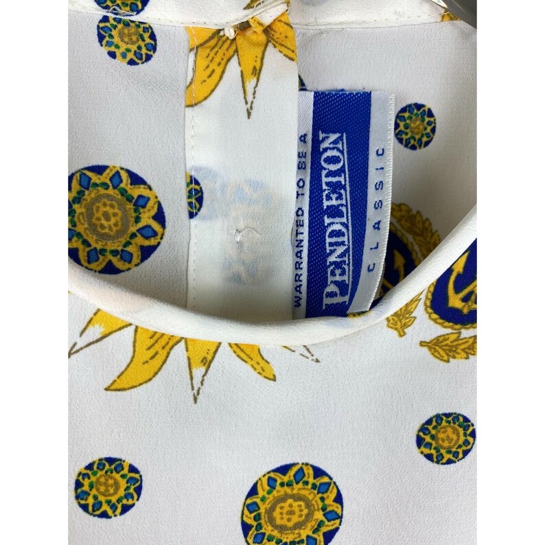 Puede incluir: Tela blanca con un estampado repetido de girasoles amarillos y dise&ntilde;os circulares azules y amarillos. Se ve una etiqueta Pendleton Classic azul y blanca. La tela parece ser parte de una camisa o blusa.
