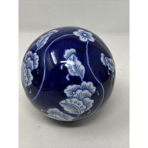 May include: A spherical ceramic decorative object featuring a deep cobalt blue glazed finish with a contrasting white hand-painted floral and vine pattern. The surface is smooth and glossy, showcasing stylized blossoms and leaves wrapping around the sphere.