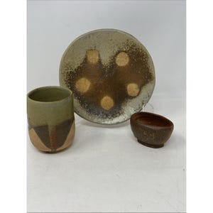 May include: A ceramic tableware set featuring a plate, a cup, and a small bowl. The plate is round with a brown and tan design. The cup is green and brown, and the bowl is brown. The set is likely handmade.