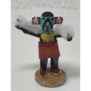 May include: A hand-painted wooden doll with a blue face, yellow and black details, and red skirt. The doll is wearing white feather headdresses and is standing on a wooden base.