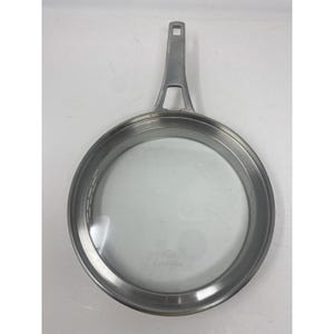 May include: A stainless steel frying pan with a clear glass lid. The pan has a long handle with a hole for hanging. The brand name "Calphalon" is visible on the lid. The pan is designed for cooking.