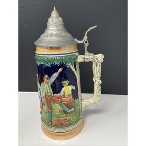 May include: A ceramic beer stein with a hinged metal lid and ornate handle. The stein features a painted scene of people dancing, with the words "Tanz auf der Alm" at the base. The colors include blue, green, yellow, and white.