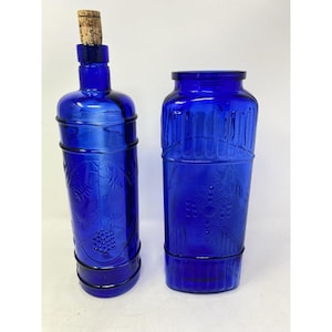 May include: Two cobalt blue glass bottles. One is a wine bottle with a cork stopper and black wire detailing. The other is a tall, cylindrical vase with a similar design. Both bottles have embossed grape and vine patterns.