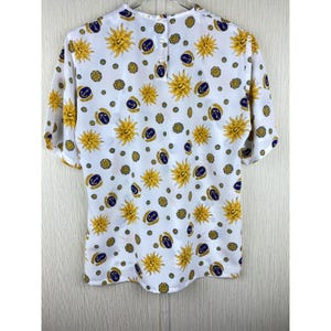 Puede incluir: Blusa blanca de manga corta con un estampado repetido de soles amarillos, anclas azul marino y c&iacute;rculos dorados. La blusa tiene un cierre de bot&oacute;n en la parte posterior del cuello y est&aacute; hecha de tela ligera.