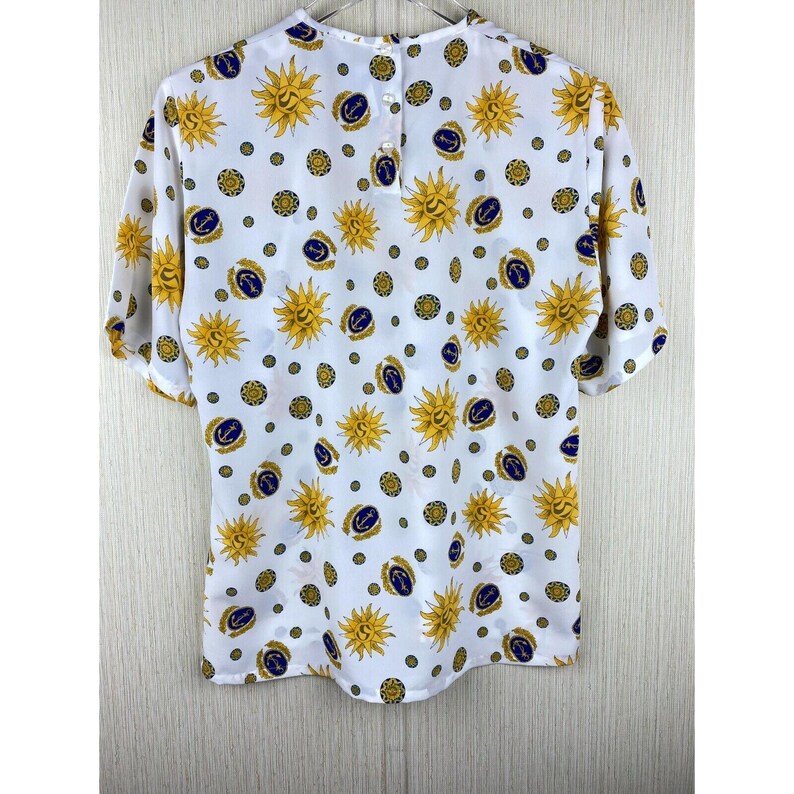 Puede incluir: Blusa blanca de manga corta con un estampado repetido de soles amarillos, anclas azul marino y c&iacute;rculos dorados. La blusa tiene un cierre de bot&oacute;n en la parte posterior del cuello y est&aacute; hecha de tela ligera.