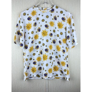 Puede incluir: Blusa blanca de manga corta con un estampado repetido de soles amarillos, anclas azul marino y medallones dorados. La blusa tiene un cuello alto y est&aacute; colgada de una percha blanca.