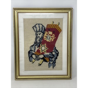 May include: Framed needlepoint art depicting a man with a long beard and hat holding a red Torah scroll. The artwork features a gold frame and is set against a beige background. The scroll has yellow and gold accents.