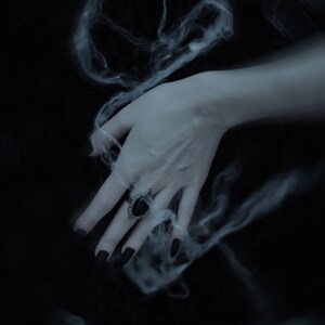 May include: A hand with black nail polish wears a silver ring with a black oval gemstone. The hand is surrounded by wispy white smoke against a dark background.
