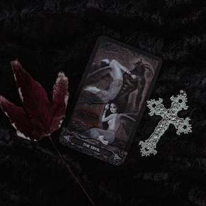 Peut inclure: Une carte de tarot représentant le Diable, dans un style gothique sombre avec une femme et une figure cornue. La carte est sur un fond noir avec une croix argentée et une feuille rouge.