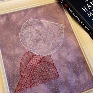 May include: A cross-stitch embroidery of a figure wearing a white bonnet and a red dress. The figure is against a purple background. The embroidery is framed in a light wood frame.