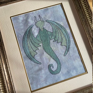 May include: A framed cross-stitch embroidery of a green dragon with wings outstretched against a light blue and purple background.