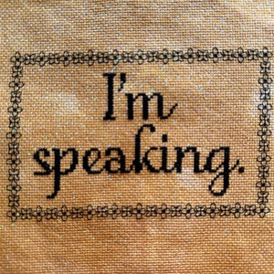 May include: Cross-stitch textile with the phrase "I'm speaking" in black script. The text is framed by a delicate floral border. The background is a tan fabric with a subtle glitter effect.