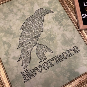 May include: A framed cross-stitch embroidery featuring a black raven with the word "Nevermore" stitched below it. The background is a light green with a tie-dye pattern.