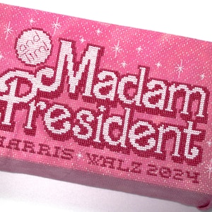 May include: Pink canvas with a white cross-stitch design that reads "Madam President" with the text "and finally" in a circle above the word "Madam". The bottom of the canvas reads "HARRIS * WALZ 2024".