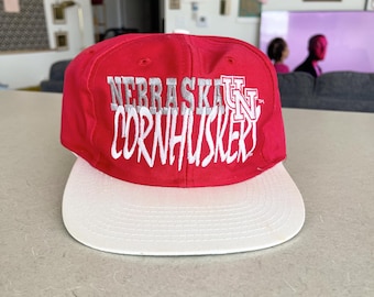 May include: A red and white baseball cap with the text "NEBRASKA CORNHUSKERS" embroidered in white and silver. The cap has a flat brim and a structured crown, perfect for sports fans.