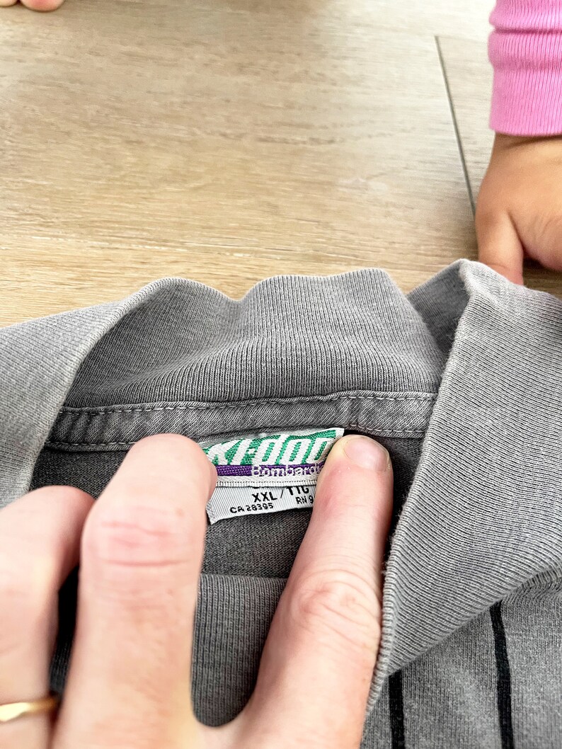 Może przedstawiać: Szara w paski koszulka z zielono-białą metką z napisem "XL/T1G Bombardir CA 28395 RN 9"